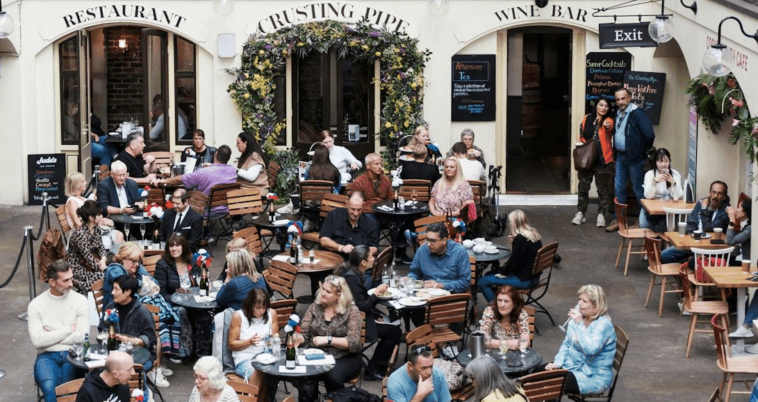Crowded outdoor courtyard with people dining at tables outside restaurants and a wine bar.